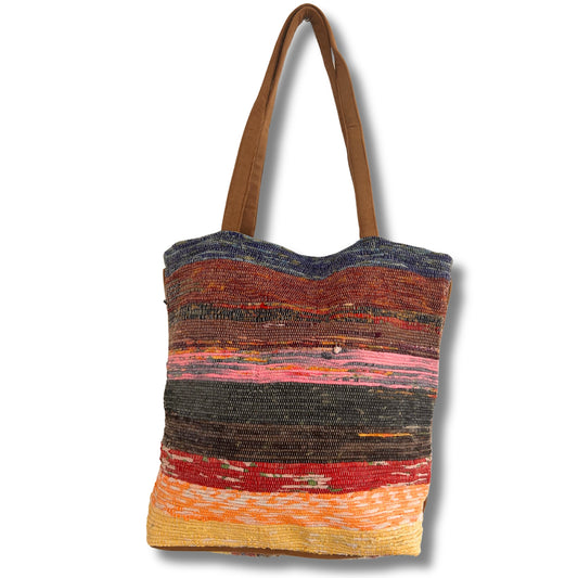 A colorful woven tote bag with brown handles. The bag features horizontal stripes in a variety of colors, including blues, reds, pinks, and yellows, set against a plain background.