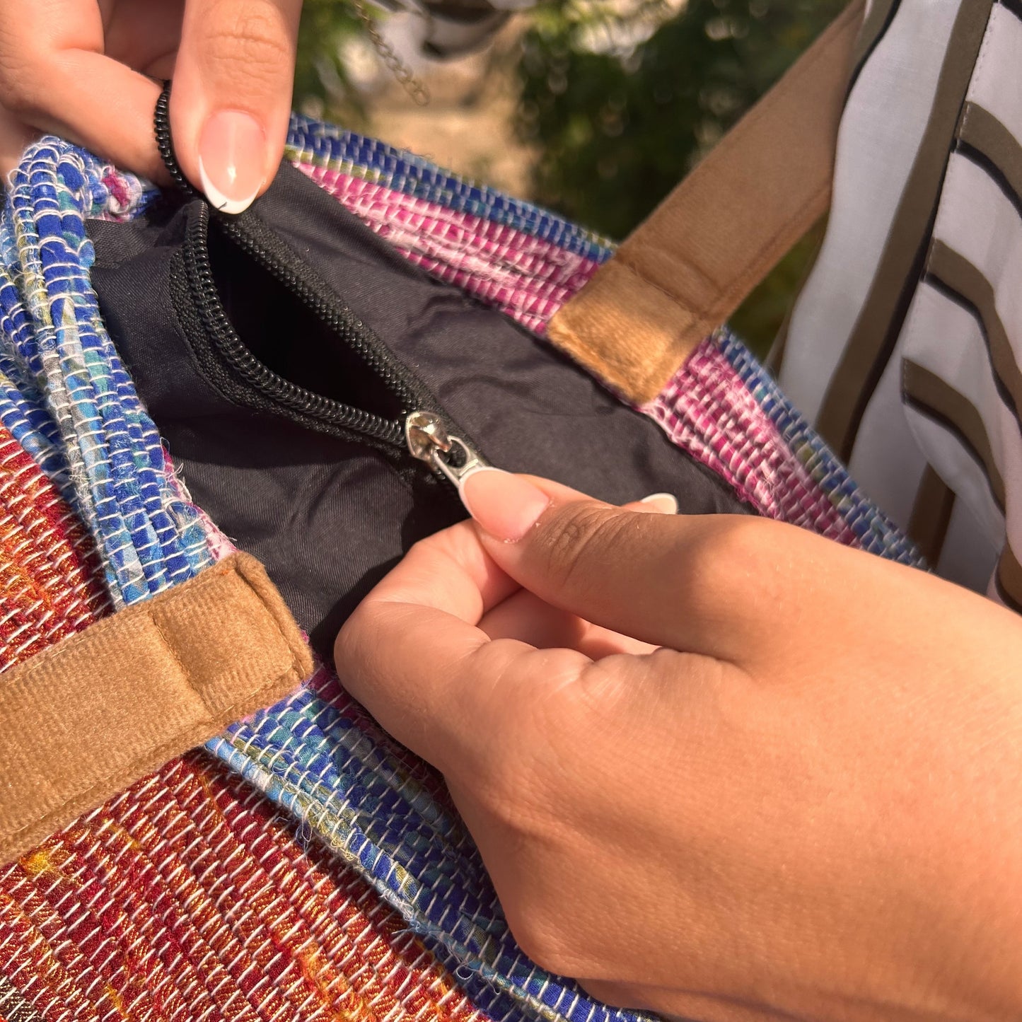 Hands open a zipper on a colorful woven bag. Sunlight illuminates the scene, with greenery and part of a striped shirt visible in the background.