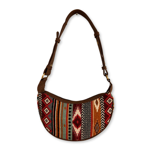 A multicolored, patterned handbag with a curved shape, featuring a brown adjustable strap. The fabric has geometric designs, creating a bohemian style against a plain white background.