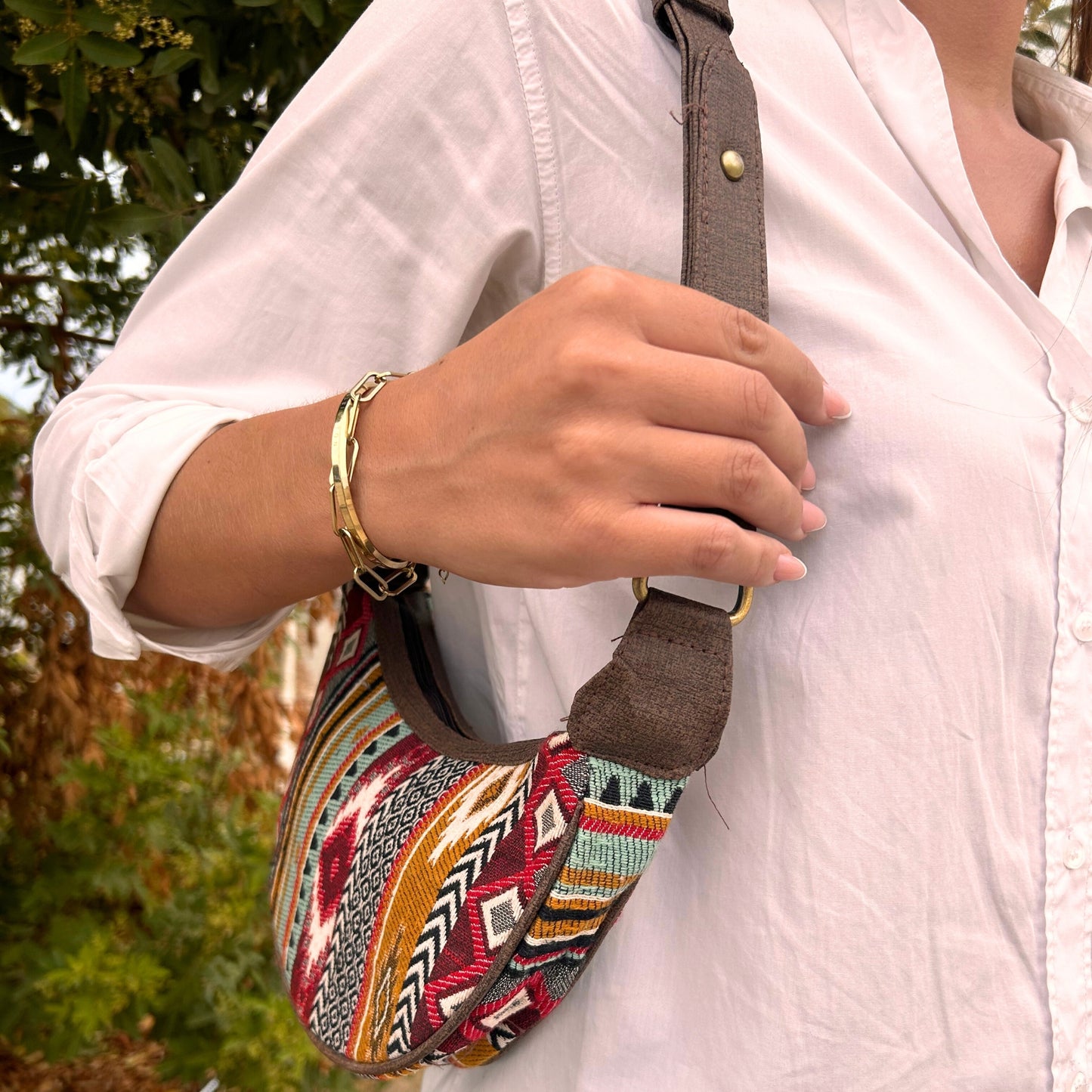 A person wearing a white shirt holds a colorful patterned bag with a brown strap and metal ring, standing outdoors near green foliage. They have a gold bracelet on their wrist.