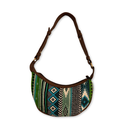 A fabric shoulder bag with a brown strap displays colorful geometric and tribal patterns in green, blue, and beige, set against a plain white background.