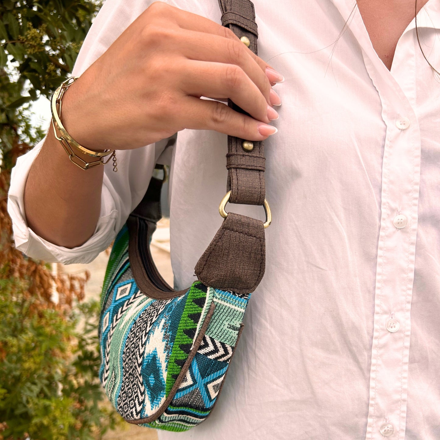 A person in a white shirt carries a small, colorful, patterned bag with a brown strap amidst green foliage. The individual wears a gold bracelet on their wrist.