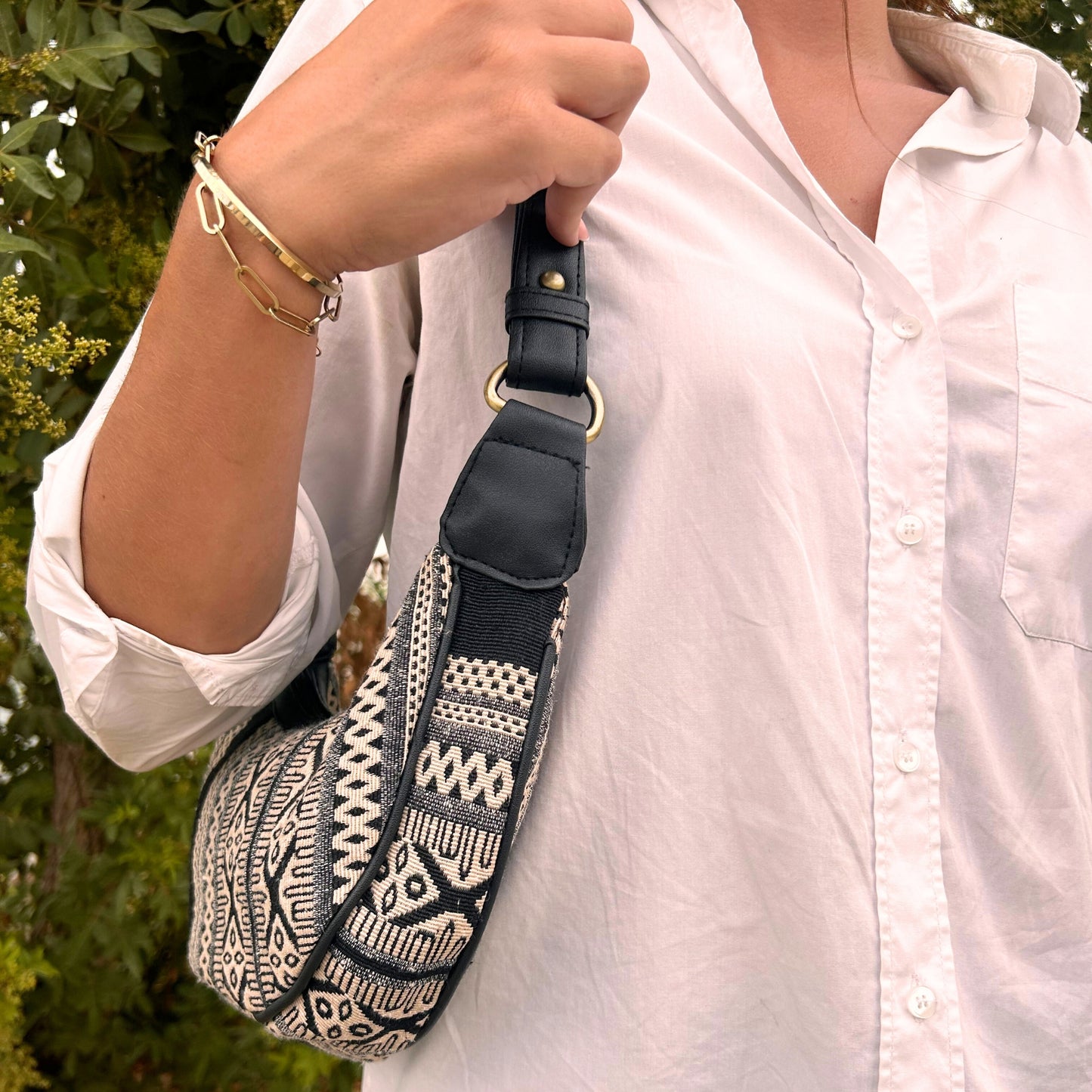 A person dressed in a white shirt holds a black and white patterned bag over their shoulder, with greenery visible in the background. They wear gold bracelets on one wrist.