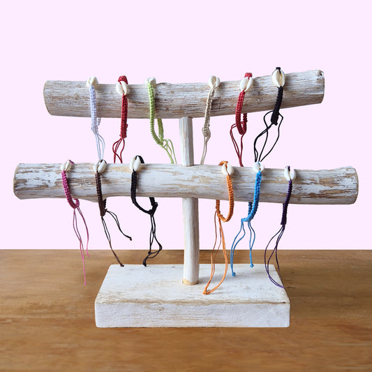 Wooden bracelet stand displaying colorful string bracelets with seashell accents, positioned on a wooden table against a plain pink background.