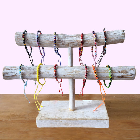 Wooden stand displaying colorful beaded bracelets hanging on two horizontal bars, placed on a light wooden surface against a pastel pink background.