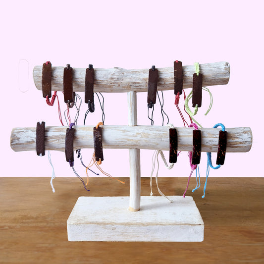 Bracelets hang on a two-tier wooden display stand. Each bracelet features brown wooden accents with colorful strings. The stand rests on a wooden surface against a pastel pink background.
