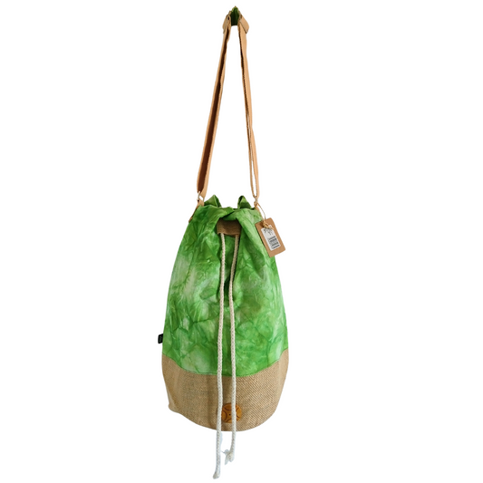 Drawstring bag with a green tie-dye fabric and beige burlap base hangs by a tan strap. A small tag is attached, displaying a tree illustration and barcode.