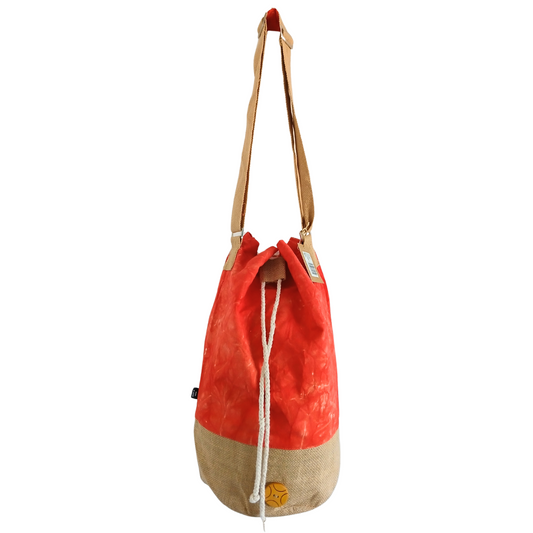 A red and beige canvas drawstring bag hangs with a brown adjustable strap. It features a rope closure and a small circular logo on the beige section. The background is plain.