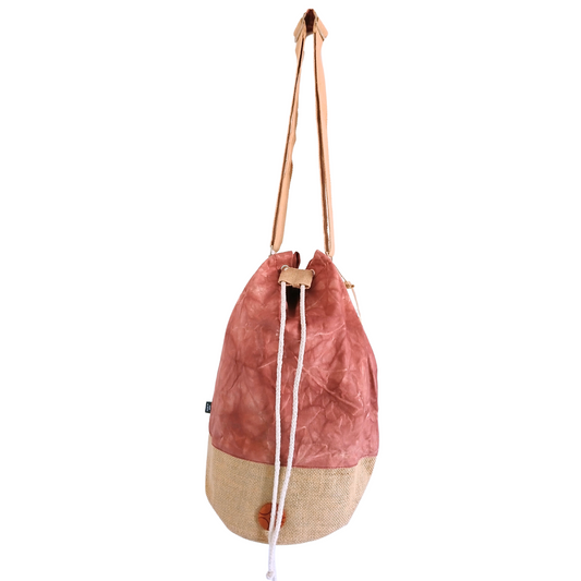 A two-tone drawstring bag hangs with a tan canvas base and a pinkish-red fabric upper, featuring long brown straps and a visible knot. The background is plain white.