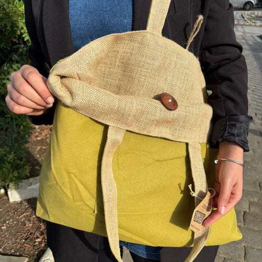 A person holds a green and burlap bag with a wooden button. A tag reads "100% Cotton." The person is outdoors, wearing a black jacket and blue shirt.