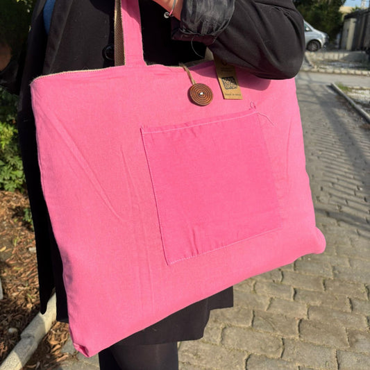 A person holds a large pink fabric tote bag with a front pocket, wooden button, and a “100% cotton made in India” label, standing on a sunlit cobblestone path.