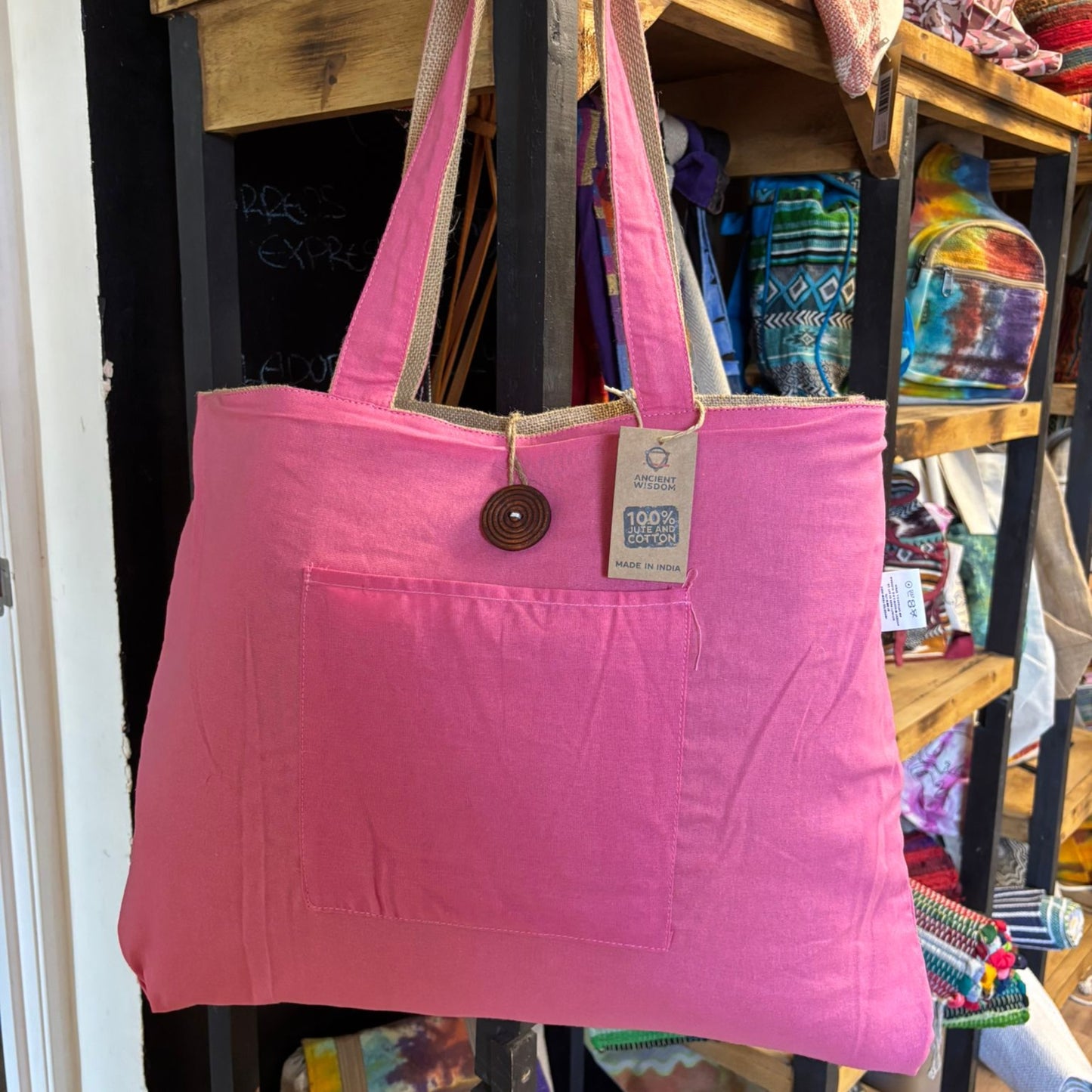 A pink tote bag hangs on display, featuring a button closure and tag reading "Ancient Wisdom, 100% Pure Cotton, Made in India," situated in a store with colorful items.
