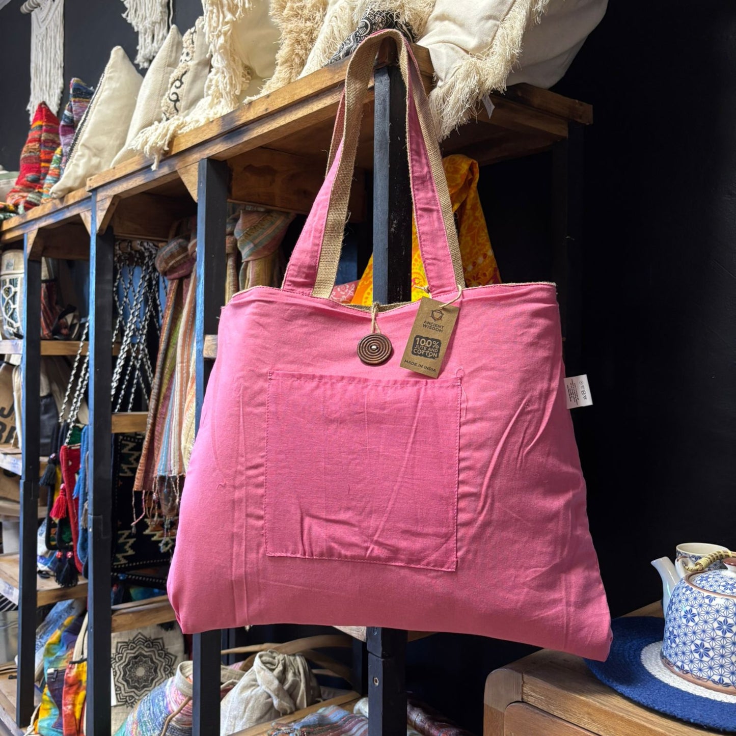 A pink fabric tote bag, with a front pocket and brown handles, hangs against wooden shelves displaying colorful textiles. Tag reads: "Lokal Made" and "100% cotton, made in Nepal."