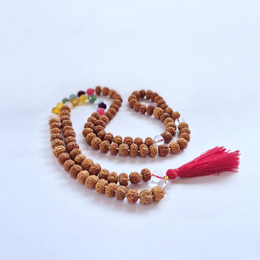 A string of rudraksha prayer beads rests on a white surface, featuring colorful round accents and ending with a vibrant red tassel.