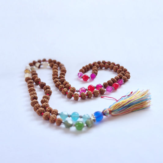 A strand of mixed beads, including brown rudraksha and colorful glass, lays coiled with a multicolored tassel attached, set against a plain white background.