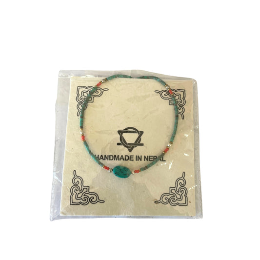 Bracelet featuring turquoise and red beads arranged in a pattern, displayed on a card with decorative corners, within clear packaging. Text reads "HANDMADE IN NEPAL" with a symbol above.