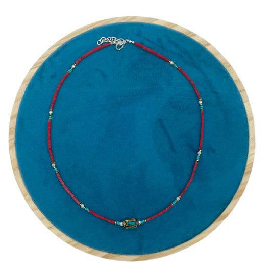 A red-beaded necklace with a cylindrical, multicolored pendant lies on a round, blue velvet surface framed by light wood.