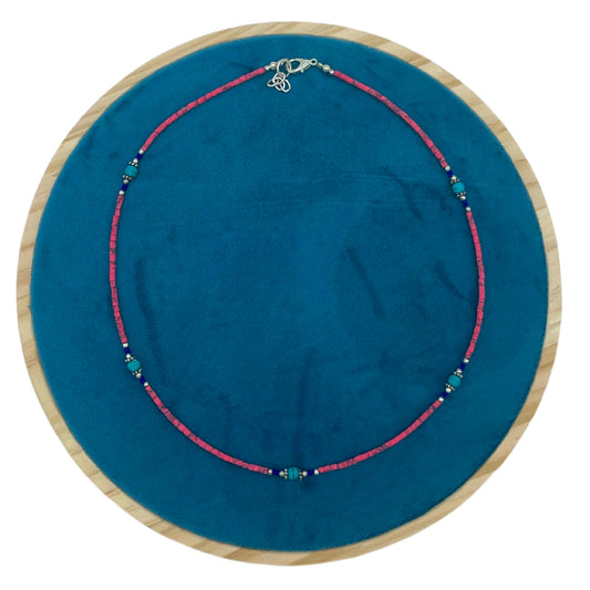 A pink beaded necklace with turquoise accents lays coiled on a blue circular fabric, surrounded by a wooden frame.