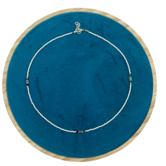 A beaded necklace lies coiled on a turquoise fabric within a round wooden frame. The necklace features small white beads interspersed with sections of colored beads.