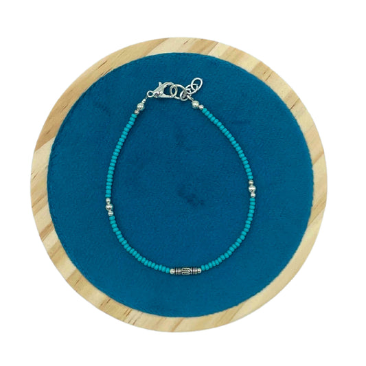 A turquoise beaded anklet with silver accents and a clasp rests on a circular blue cloth background, bordered by a wooden ring.