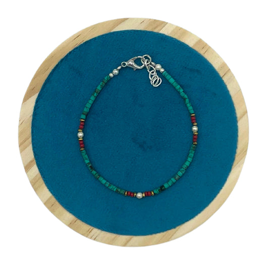 A beaded bracelet, with turquoise and red beads and silver clasp, rests on a round blue fabric inside a wooden frame.