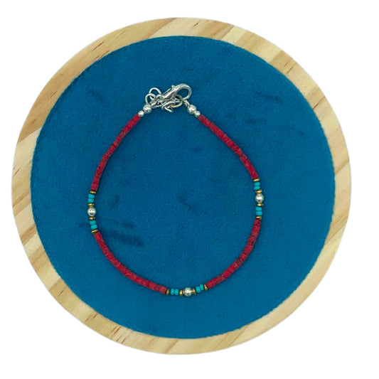 A red beaded bracelet with turquoise and gold accents rests in a circular pattern, placed on a blue fabric within a wooden frame.
