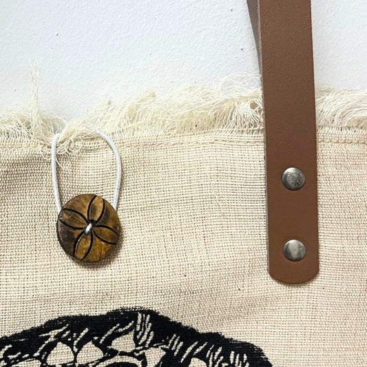 A round, decorated button with a floral pattern hangs on a string from a fringed beige fabric. It is next to a brown leather strap with metal rivets, set against a textured wall.