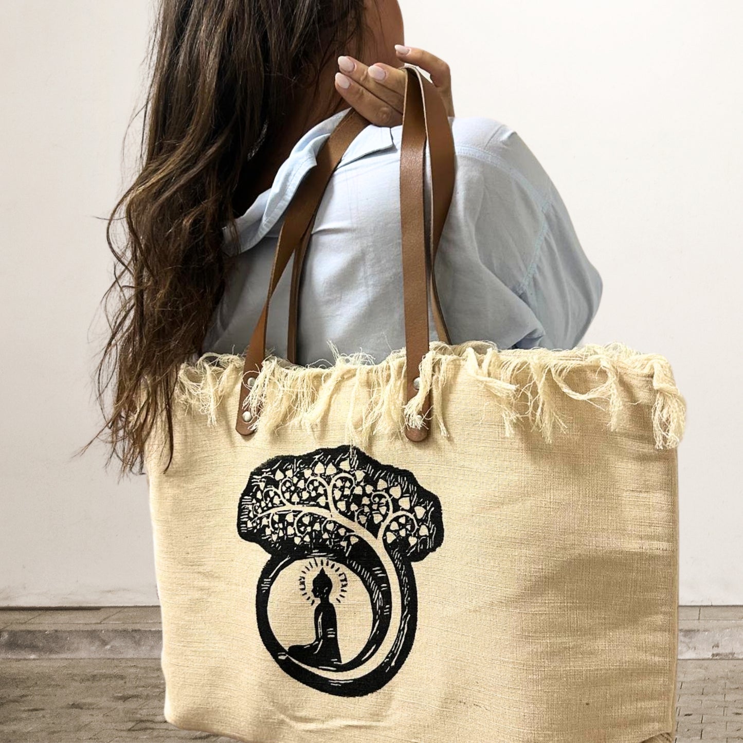 A beige tote bag with a fringe top shows a silhouette of a meditating figure under a tree. The person, wearing a light blue shirt, carries it in an indoor setting.