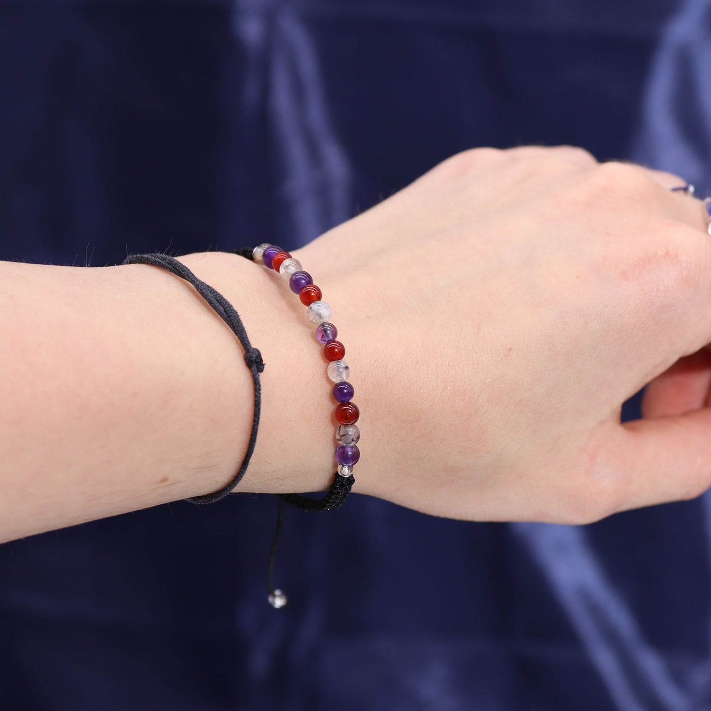 A wrist is adorned with two bracelets: one featuring multicolored beads and the other a simple black string. The background is a smooth, dark blue fabric.
