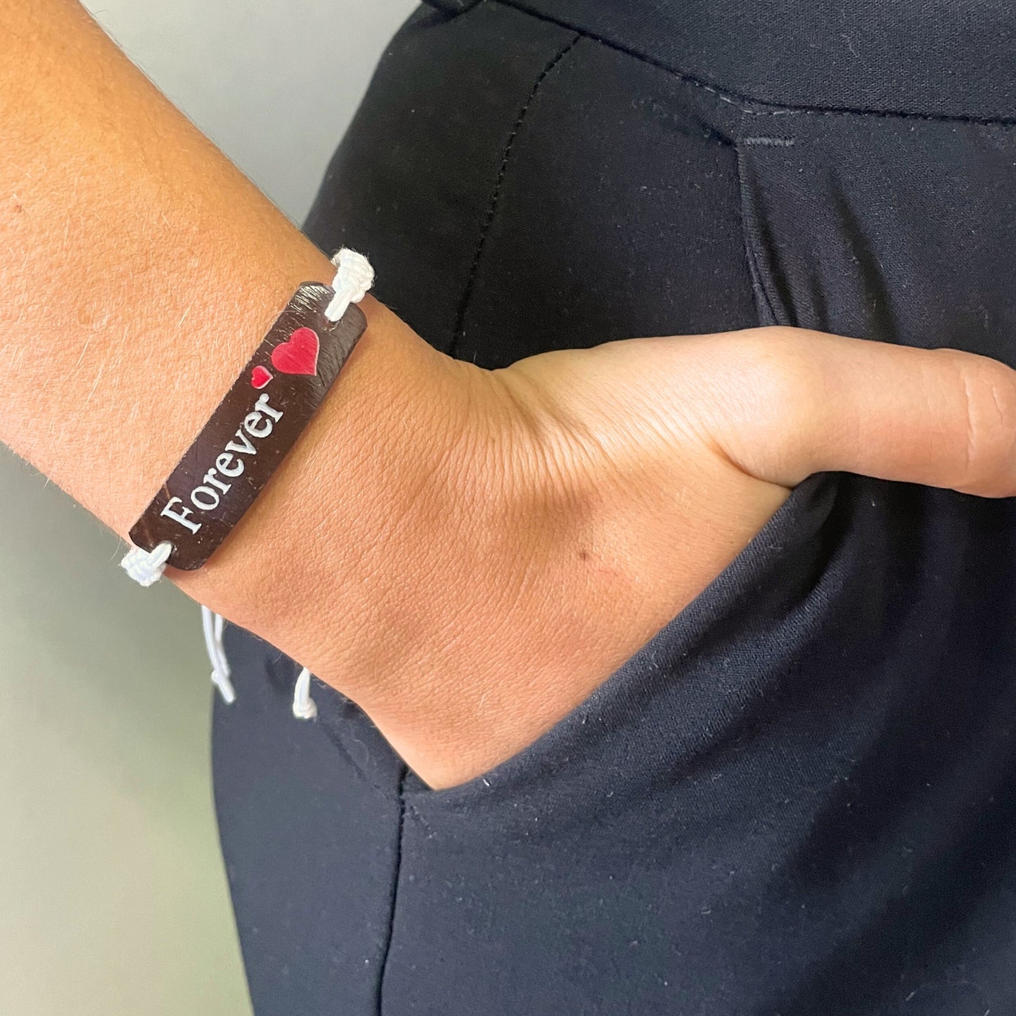 A wrist sports a bracelet with the text "Forever" and a red heart, as the hand rests in a pocket against a dark fabric background.