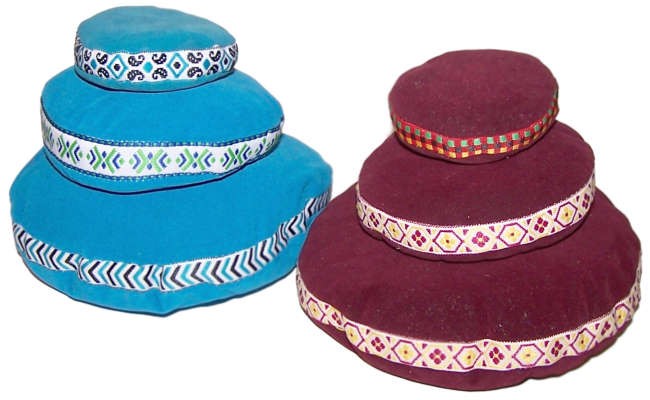 Two stacks of cushion-like poufs with decorative patterns are placed side by side. The left stack is blue with white and green trims, while the right stack is maroon with colorful trims.