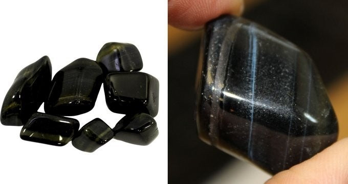 Polished, dark stones are on a white surface, seven in a pile. A single stone is held against a neutral background with a finger.
