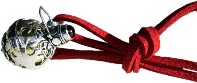 A decorative, silver-colored locket hangs from a tied red suede cord. The locket has cut-out designs and a small red stone. The backdrop is white.
