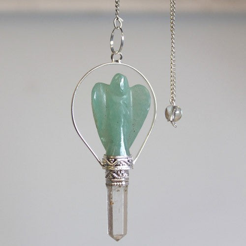 A pendulum with a green, carved-stone angel figure is suspended from a silver chain. The angel is attached to a clear crystal point, which sits against a pale gray backdrop.
