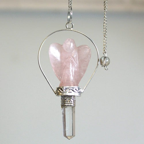 A pink, angel-shaped pendulum hangs suspended in a metal frame from a silver chain, ending in a small glass bead, against a soft, blurred background.