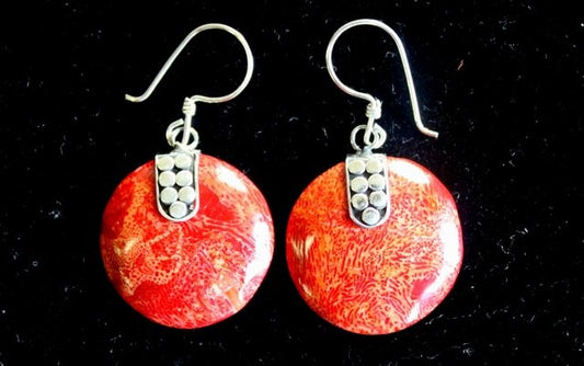 Two circular earrings dangle, set with a silver mount decorated with a cluster of small dots, with a red coral texture and a dark background.