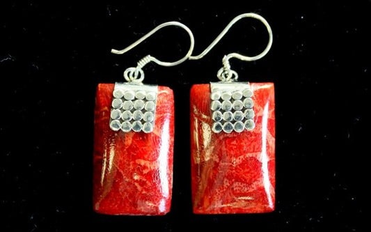 Two red, rectangular coral earrings are on display. The earrings have silver trim with ball details, and silver hooks, and they lie on a black background.