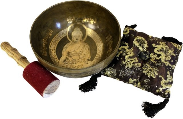 A singing bowl rests beside a wooden mallet with a red suede end. The bowl features an engraved Buddha and surrounding text, situated next to a decorative cushion with tassels.