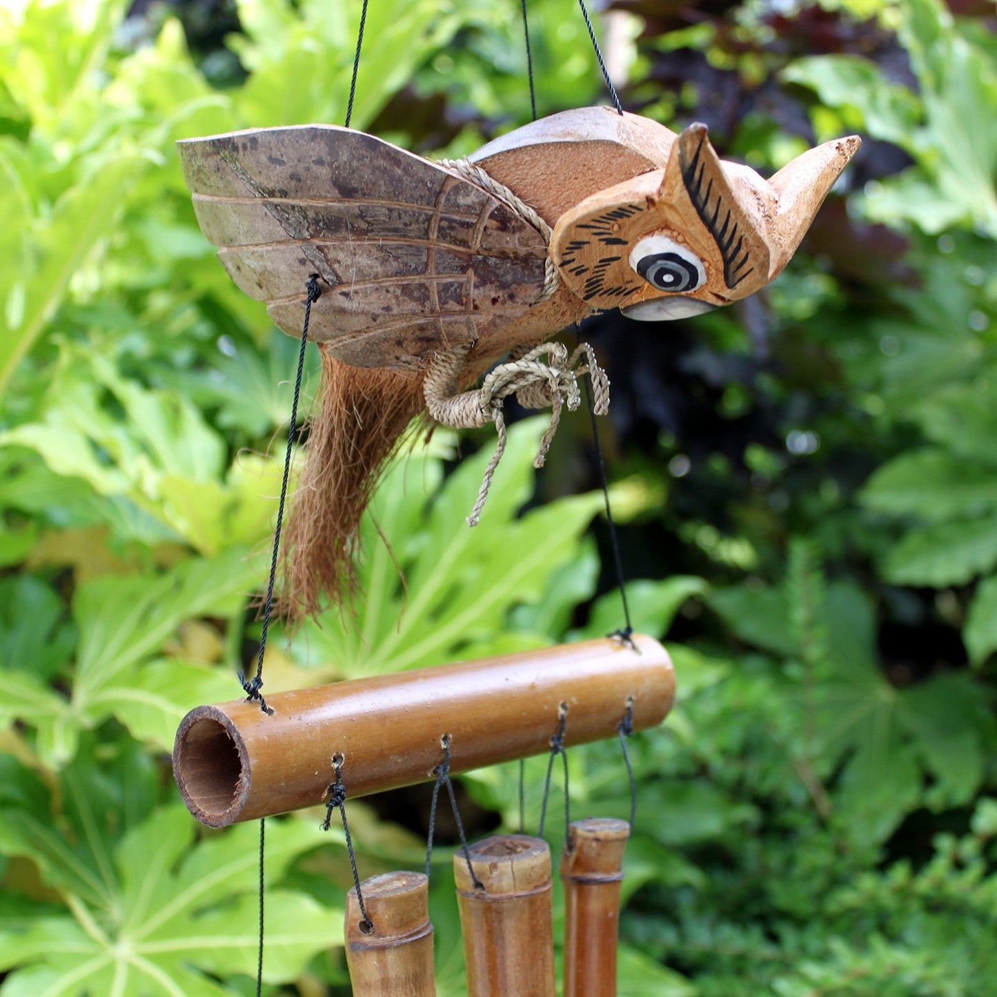 A coconut shell owl wind chime hangs amidst lush green foliage. The owl, crafted from coconut shell and wood, is attached to bamboo tubes that gently chime in the breeze.