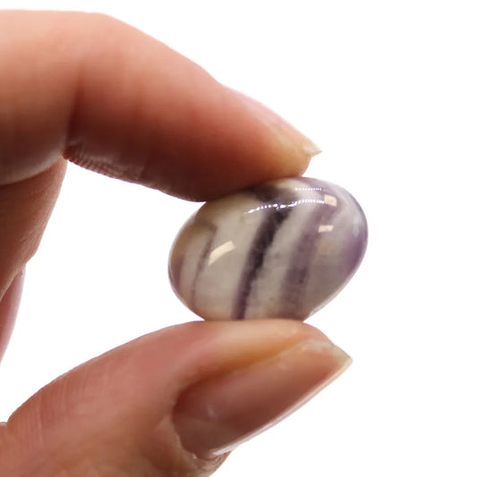 A polished, oval, striped stone is held between a thumb and forefinger against a white backdrop.