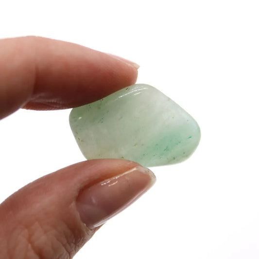 Fingers hold a polished, pale green stone. The gemstone is rounded and appears smooth, showcased against a white background.