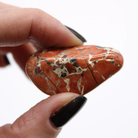 A hand with black nail polish pinches a polished, heart-shaped, reddish-orange stone with dark lines and white/gray mottling on a white background.
