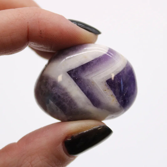 A polished stone with alternating white and purple chevrons is being held between two fingers with black nail polish against a bright white background.