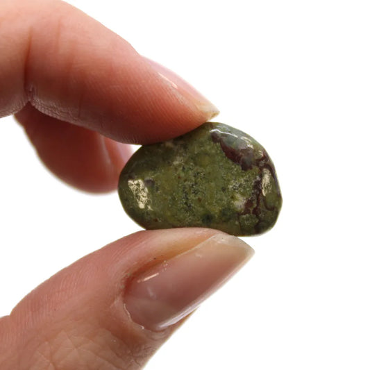 A thumb and forefinger pinch a small, polished, green rock with some reddish-brown speckles. The background is plain white.
