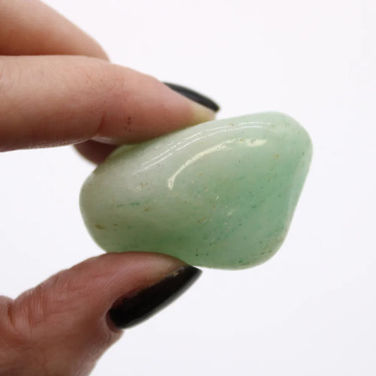 A light green tumbled stone is pinched between two fingers with black nail polish, in front of a white background.