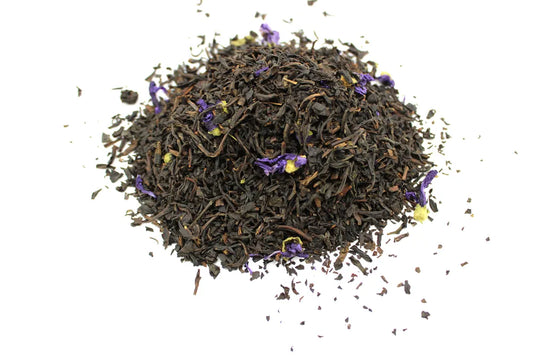 A pile of loose black tea leaves is mixed with small purple flowers, resting on a plain white surface.