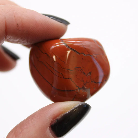A red jasper stone is being held by a person's fingertips against a white background. The fingernails are painted black.