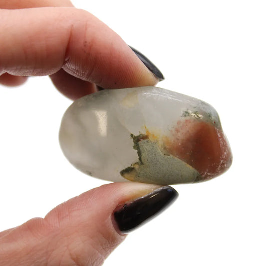 A small polished rock with a clear body, with hints of green and red inclusions, is being held between two fingers. The backdrop is plain white.