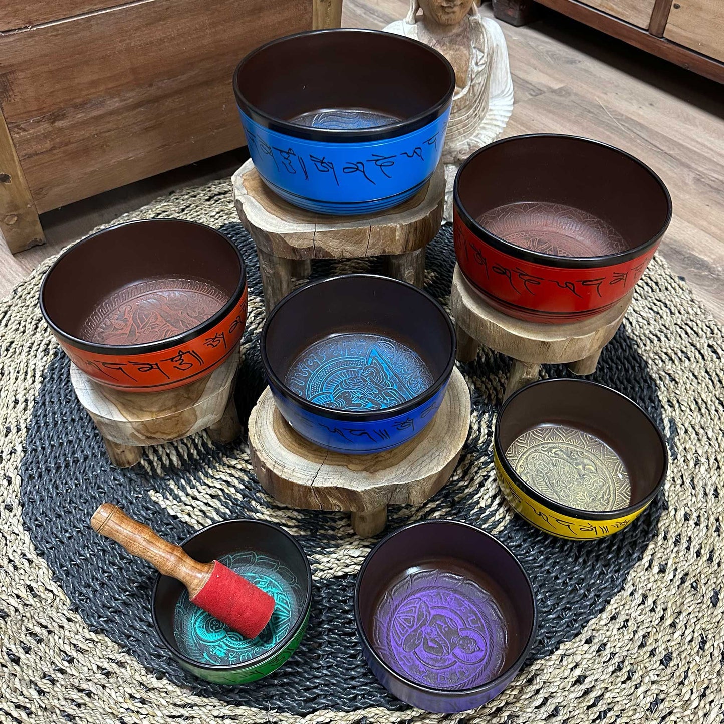 Seven colorful Tibetan singing bowls arranged on wooden stands and a woven mat with a mallet, some inscribed with ཨོཾ་མ་ཎི་པདྨེ་ཧཱུྃ (om mani padme hum).