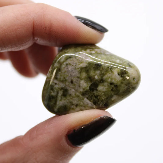 A moss-colored, polished stone is held between fingers with black polished nails against a white background.